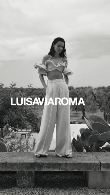 Black and white fashion shot of a young woman in a crop top and linen pants standing on a stone ledge in a rural outdoor setting.