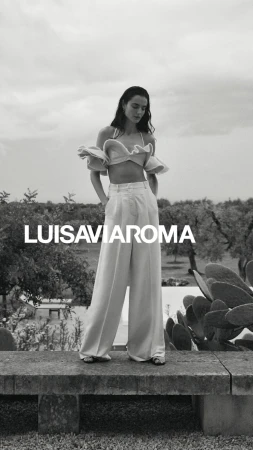 Black and white fashion shot of a young woman in a crop top and linen pants standing on a stone ledge in a rural outdoor setting.