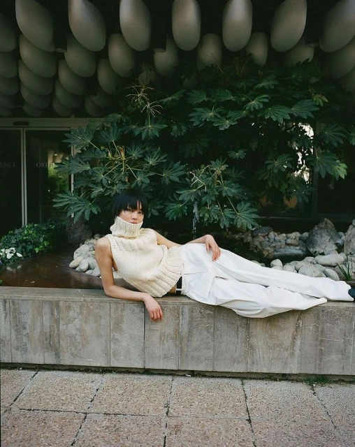 Woman in white sleeveless sweater and pants lies on concrete near greenery, with unique architectural detail overhead in an outdoor setting.