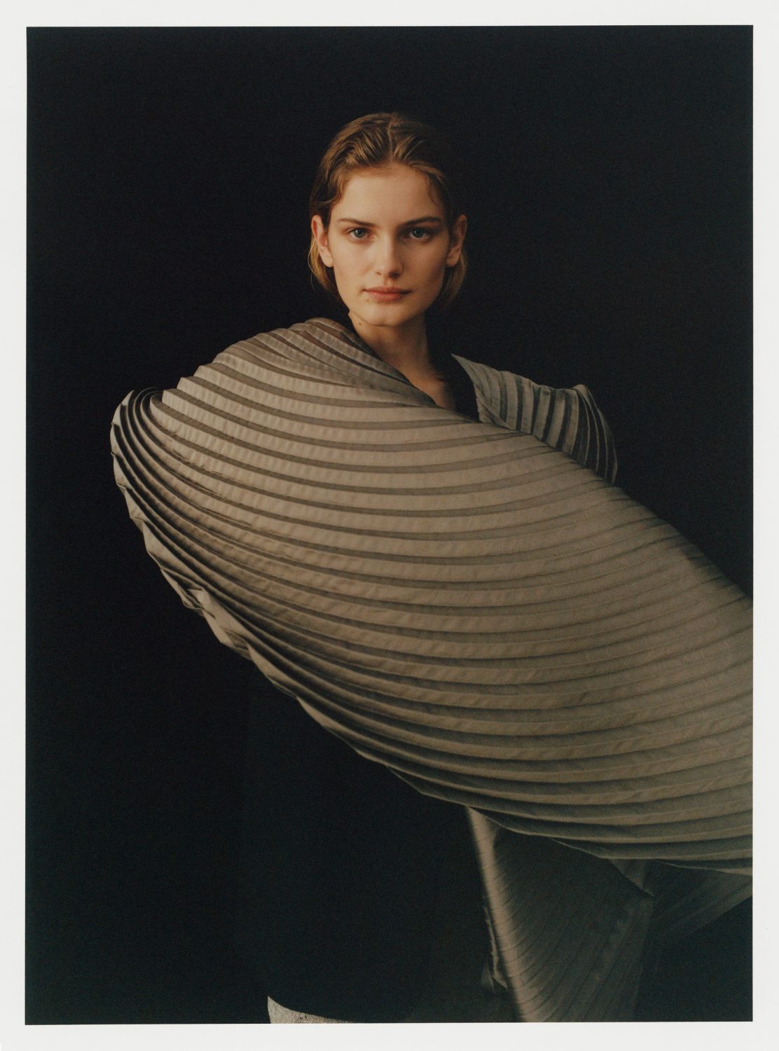 Portrait of a woman wearing a large pleated grey garment against a plain dark background.