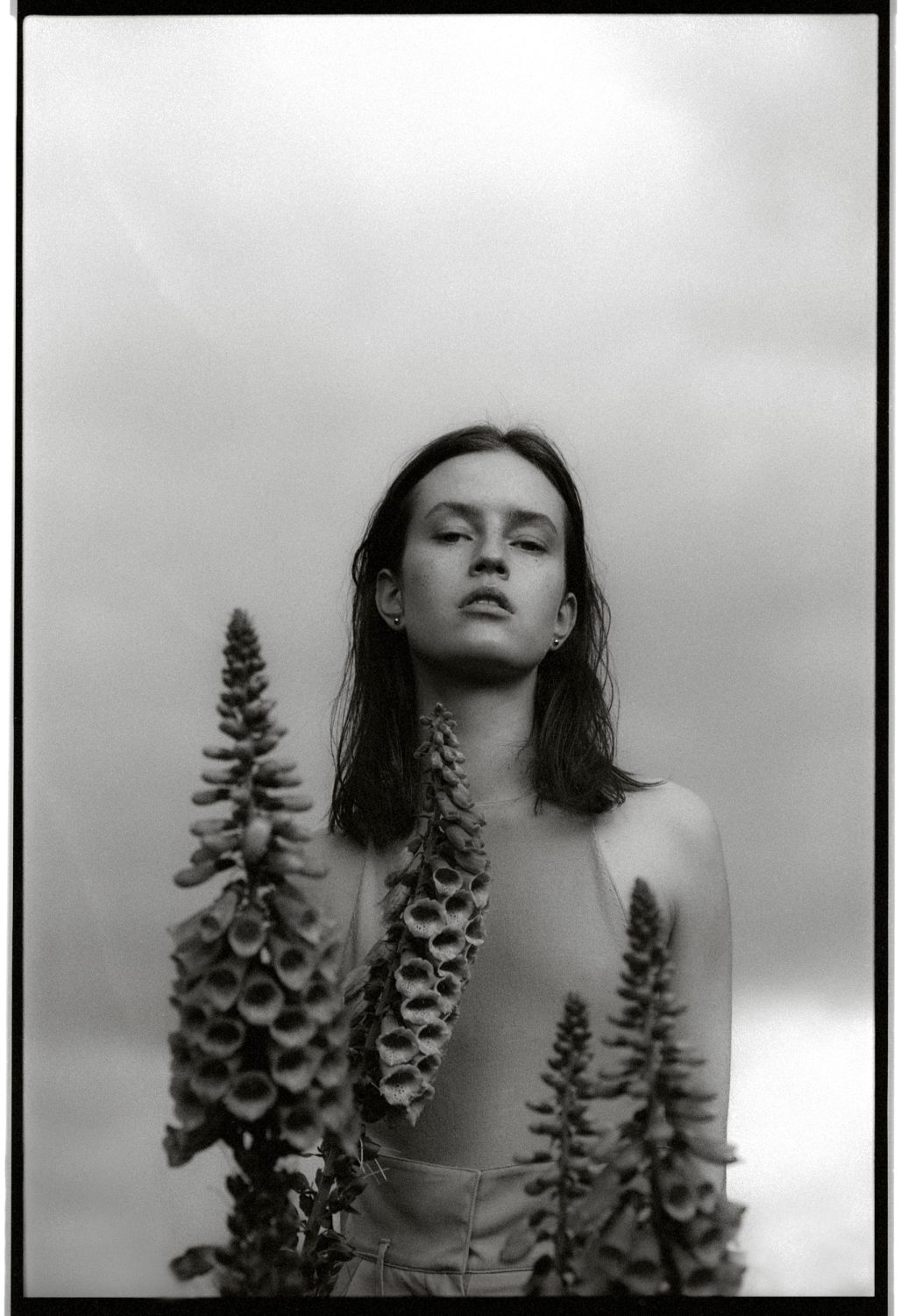 Monochrome portrait of a woman with foxglove flowers against a cloudy sky, creating a high-contrast, artistic composition.