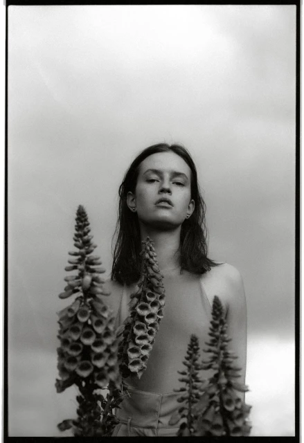 Monochrome portrait of a woman with foxglove flowers against a cloudy sky, creating a high-contrast, artistic composition.