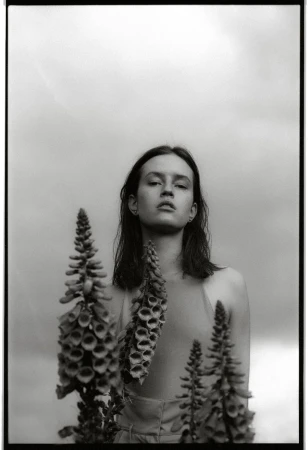 Monochrome portrait of a woman with foxglove flowers against a cloudy sky, creating a high-contrast, artistic composition.