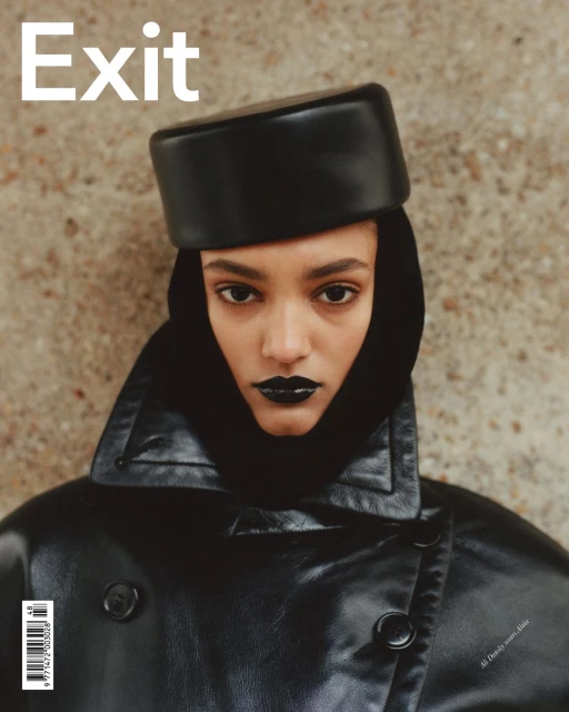 Magazine cover featuring a model with dark lipstick wearing a black leather coat and hat against a textured beige background.