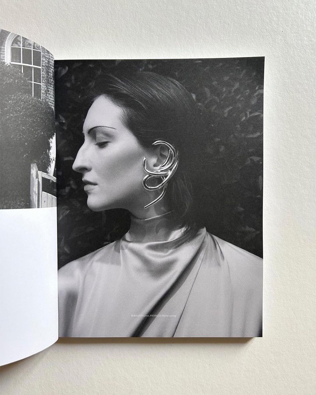 Black and white portrait of an adult woman with a sculptural metal earring, in front of a leafy background, with a book visible.