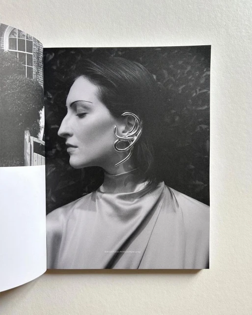 Black and white portrait of an adult woman with a sculptural metal earring, in front of a leafy background, with a book visible.