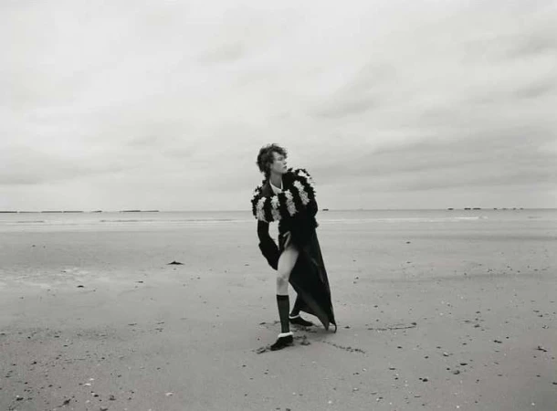Model in ruffled dark coat and knee-high socks posing on a wide sandy beach under an overcast sky by Kulesza and Pik.