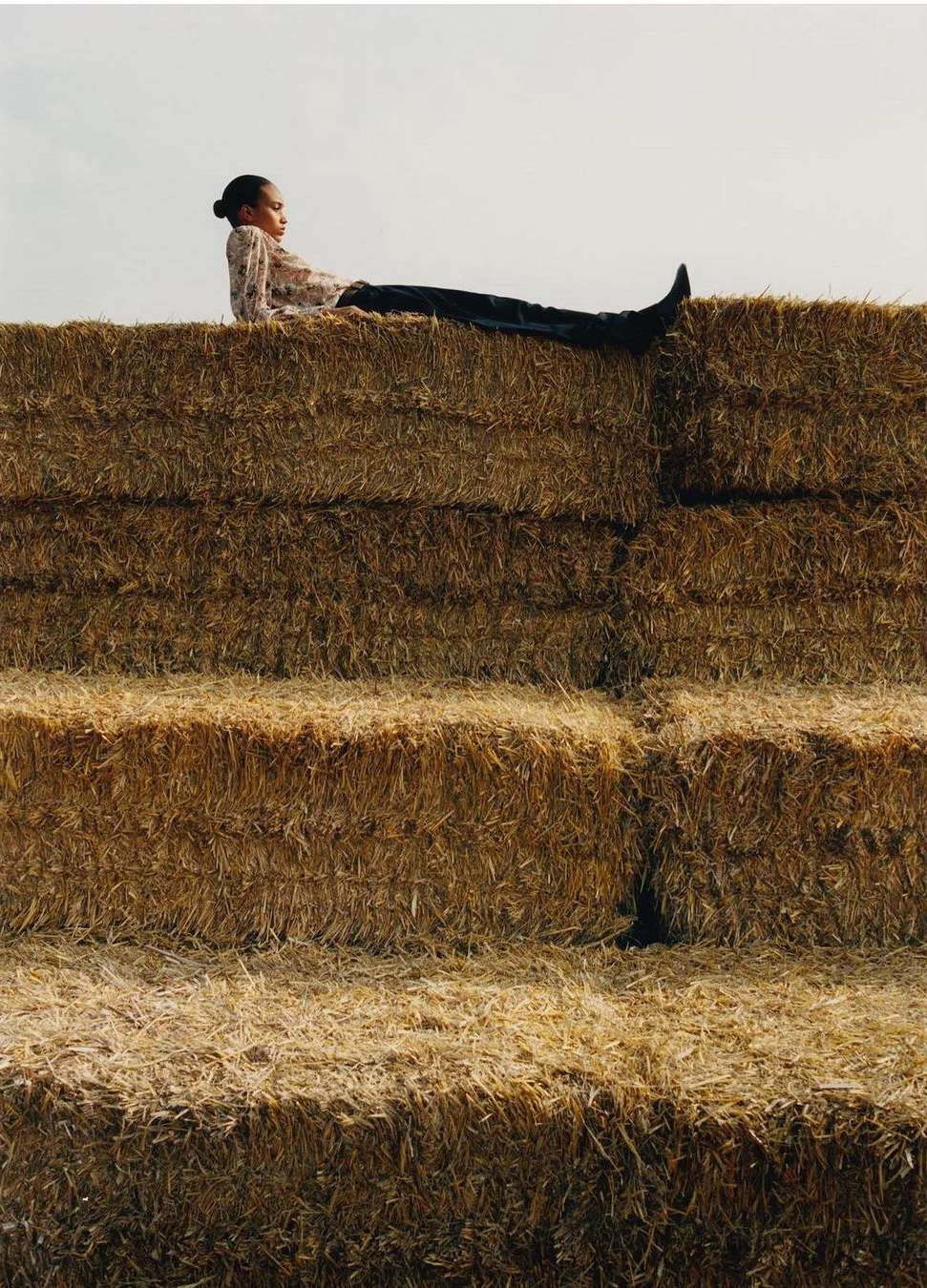 Woman in floral top and jeans sitting atop stacked hay bales in a rural outdoor setting with natural light.