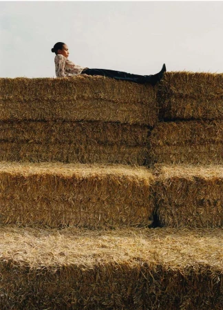 Woman in floral top and jeans sitting atop stacked hay bales in a rural outdoor setting with natural light.