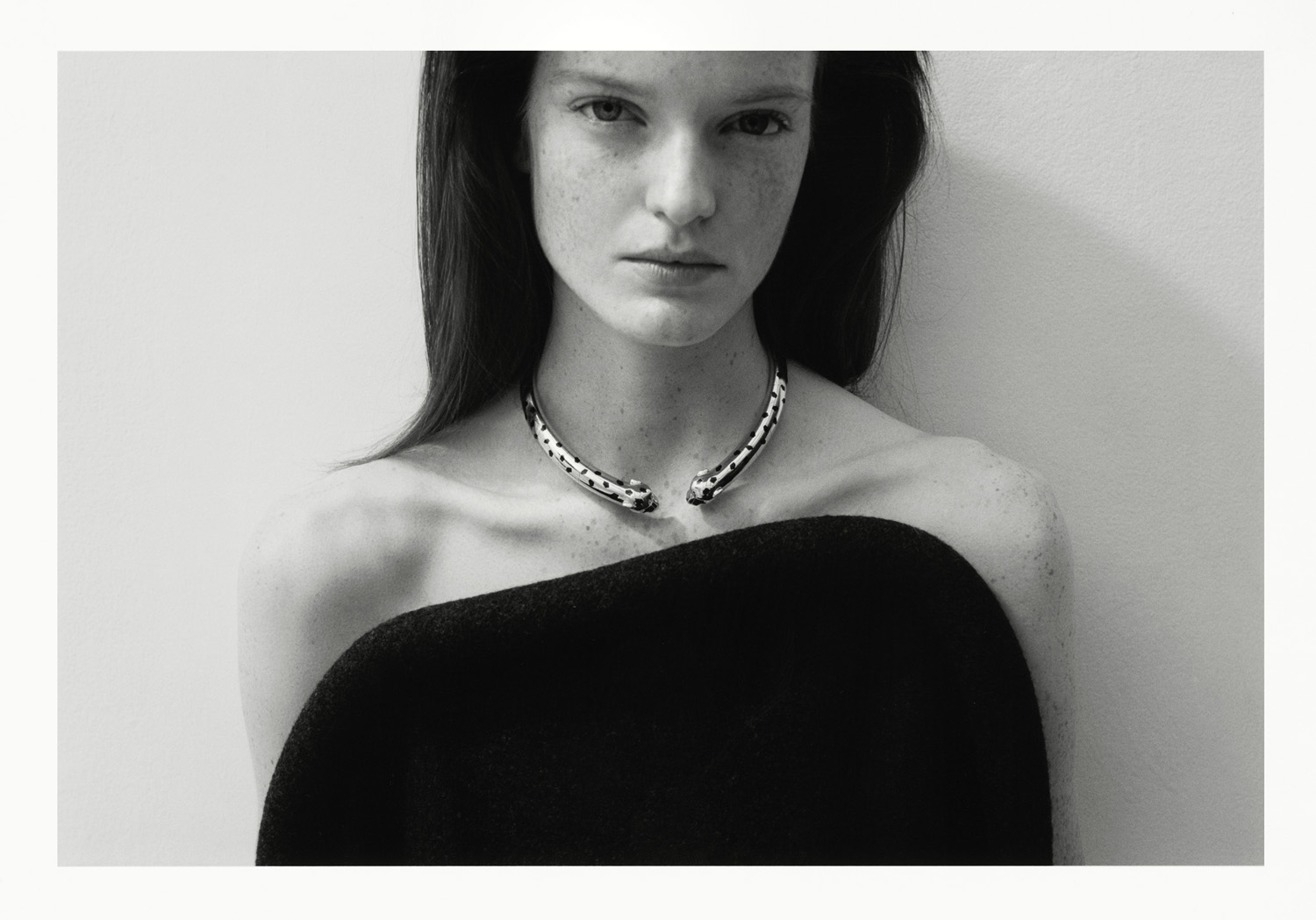 Monochrome portrait of an adult woman with dark hair, wearing a black top and silver necklace, in front of a neutral wall. Fashion photography.
