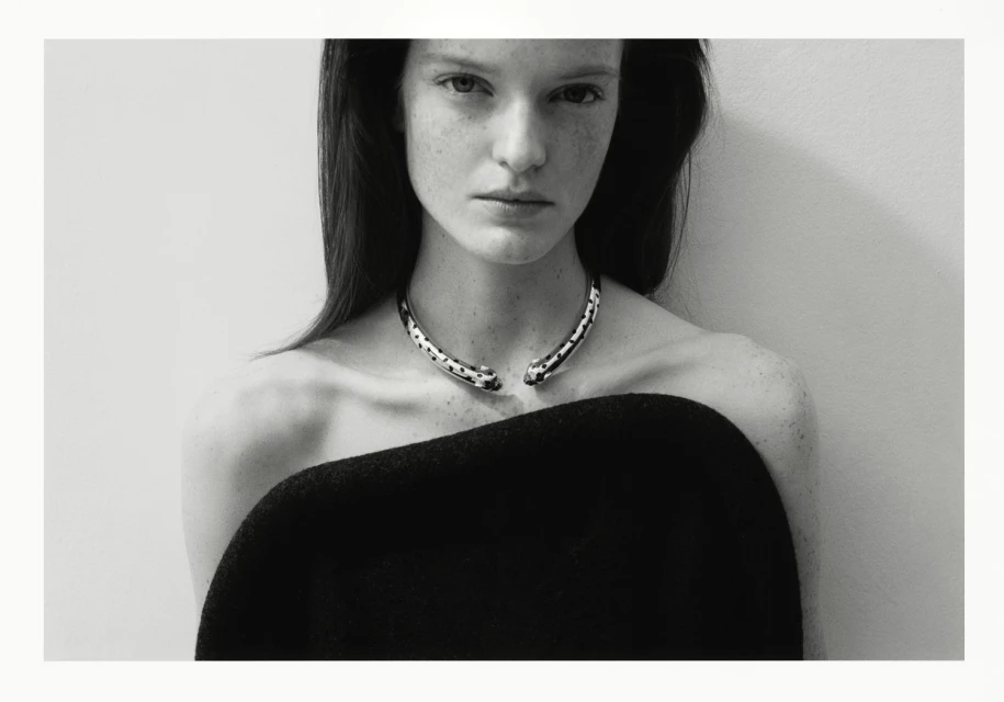 Monochrome portrait of an adult woman with dark hair, wearing a black top and silver necklace, in front of a neutral wall. Fashion photography.