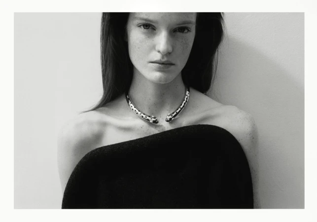 Monochrome portrait of an adult woman with dark hair, wearing a black top and silver necklace, in front of a neutral wall. Fashion photography.
