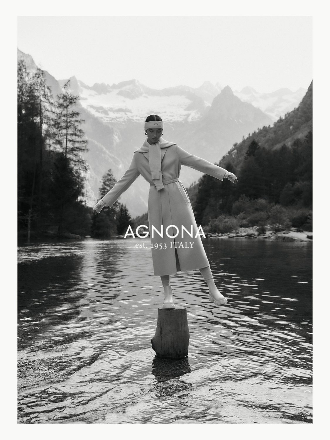 Agnona fashion editorial featuring a woman in a tan coat balancing on a tree stump in a lake with a mountain range backdrop.