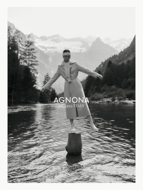 Agnona fashion editorial featuring a woman in a tan coat balancing on a tree stump in a lake with a mountain range backdrop.