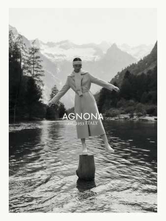 Agnona fashion editorial featuring a woman in a tan coat balancing on a tree stump in a lake with a mountain range backdrop.
