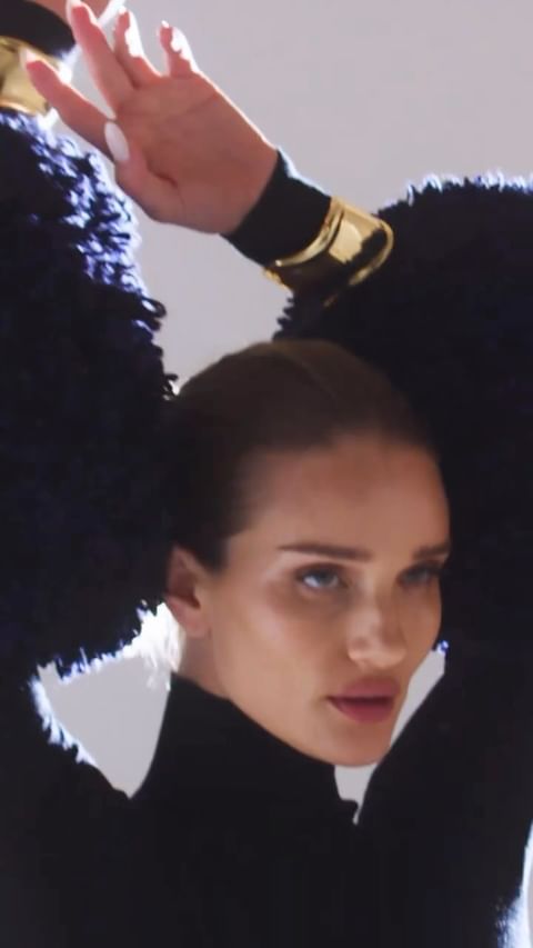 Close-up studio shot of a fair-skinned adult female model posing with her arms raised, wearing a black turtleneck and gold bracelets.