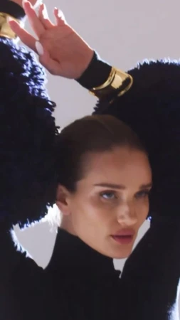 Close-up studio shot of a fair-skinned adult female model posing with her arms raised, wearing a black turtleneck and gold bracelets.