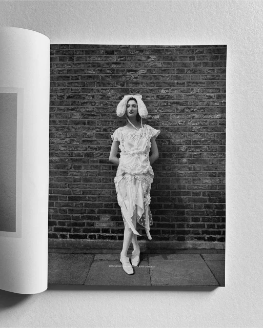 A woman in a white gown and bonnet poses against a brick wall in black and white, showcasing a fashion editorial shot.