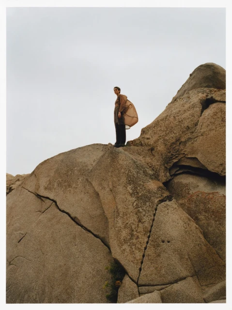 Adult female standing on a rock formation wearing a long jacket and pants, under an overcast sky in a natural environment.