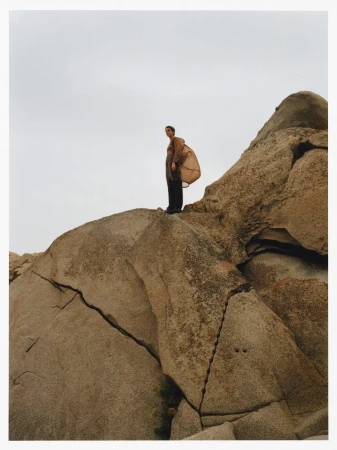 Adult female standing on a rock formation wearing a long jacket and pants, under an overcast sky in a natural environment.