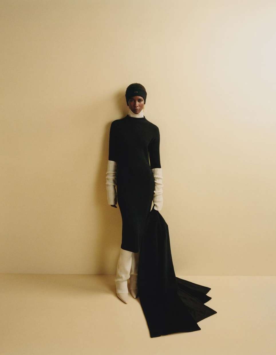 Elegant woman in a black dress, cream boots, and matching long gloves poses against a neutral backdrop in a fashion studio shot.