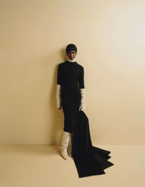 Elegant woman in a black dress, cream boots, and matching long gloves poses against a neutral backdrop in a fashion studio shot.