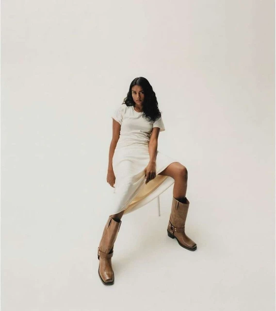 A woman in a white dress and cowboy boots poses in a studio, showcasing a fashion-forward look against a neutral backdrop.
