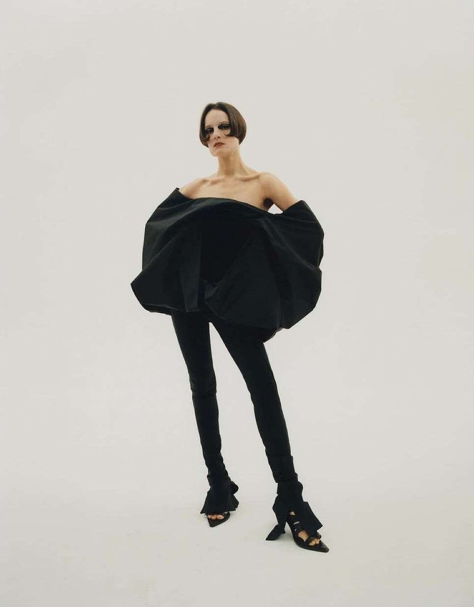 A model poses in a studio with a black, off-the-shoulder top and black leggings. She is standing in the center with a light backdrop.