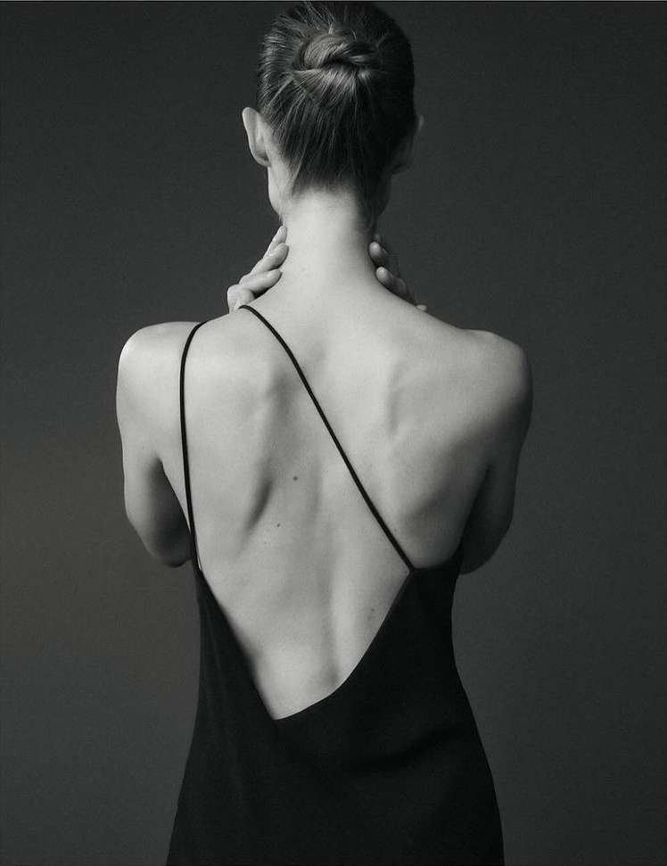 A woman with her hair in a bun wears a black dress and touches her neck in a studio shot, highlighting fashion and beauty.
