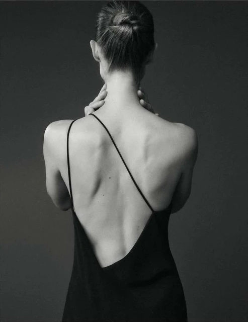 A woman with her hair in a bun wears a black dress and touches her neck in a studio shot, highlighting fashion and beauty.
