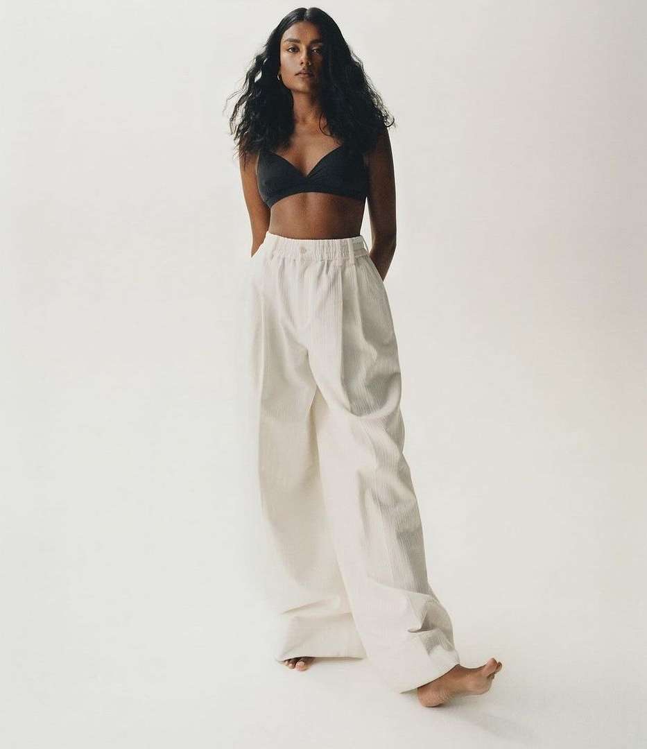 Brunette woman in black bra and white pants poses in a full shot against a white backdrop in a fashion or beauty context. 