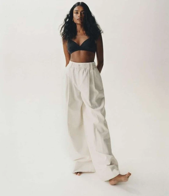 Brunette woman in black bra and white pants poses in a full shot against a white backdrop in a fashion or beauty context. 