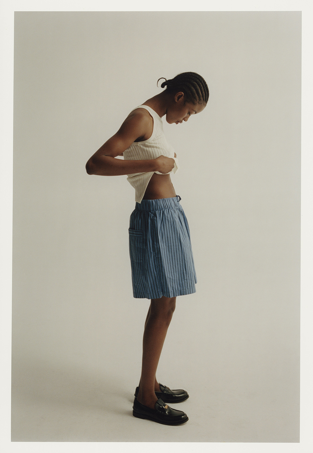 Fashion portrait of a person with cornrows adjusting their beige ribbed top in a light studio wearing pinstripe shorts and loafers.
