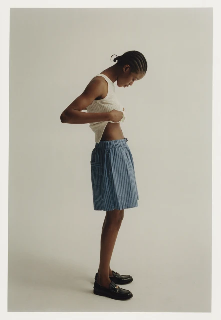 Fashion portrait of a person with cornrows adjusting their beige ribbed top in a light studio wearing pinstripe shorts and loafers.