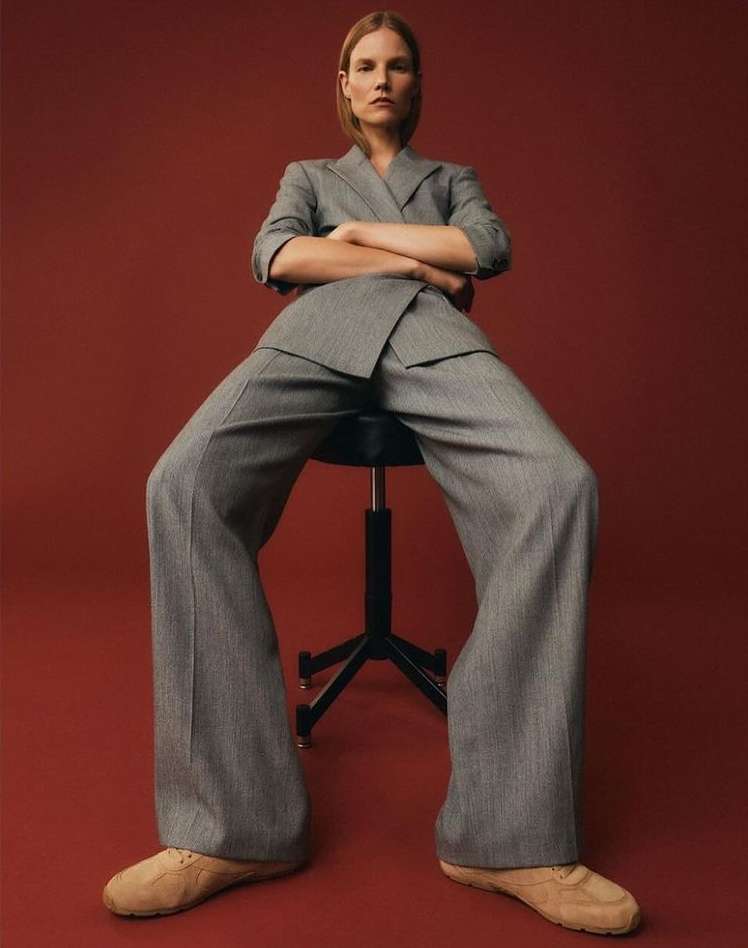 An adult female model in a grey suit poses on a black stool against a red background in a fashion forward eye-level full shot.