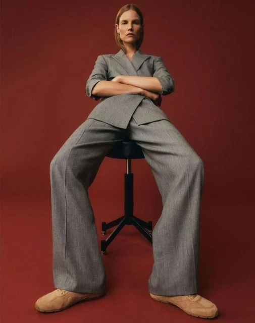 An adult female model in a grey suit poses on a black stool against a red background in a fashion forward eye-level full shot.