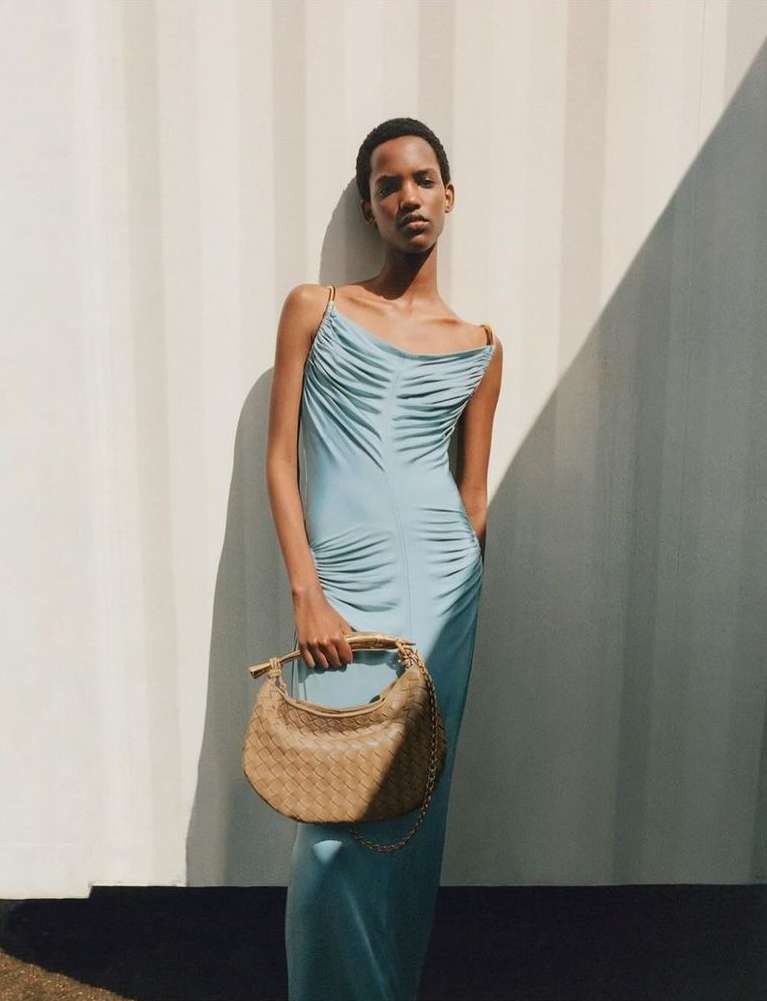 An adult female fashion model poses in a light blue dress holding a woven handbag against a wall casting a geometric shadow.