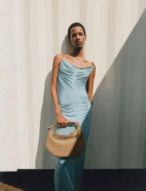An adult female fashion model poses in a light blue dress holding a woven handbag against a wall casting a geometric shadow.