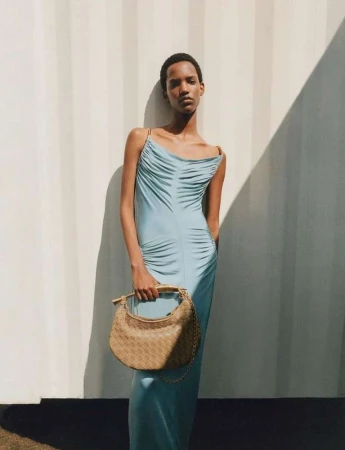 Model wearing a light blue slip dress holding a woven tan handbag against a white wall with harsh shadows