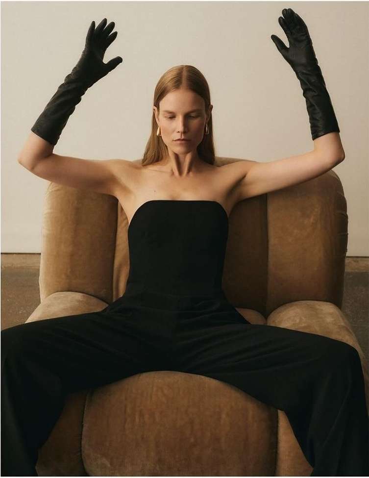Woman in black strapless jumpsuit and long leather gloves seated in an armchair with arms raised.