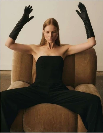 Woman in black strapless jumpsuit and long leather gloves seated in an armchair with arms raised.