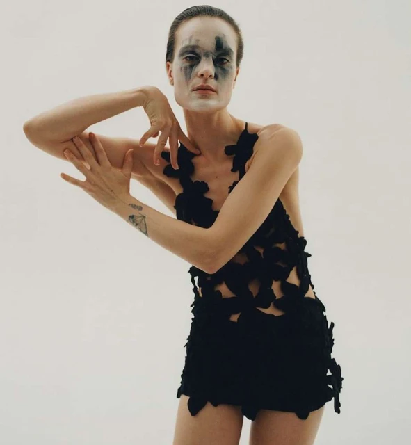 Woman poses in a dark, flower-detailed outfit against a plain white backdrop, face adorned with artistic black and white makeup.