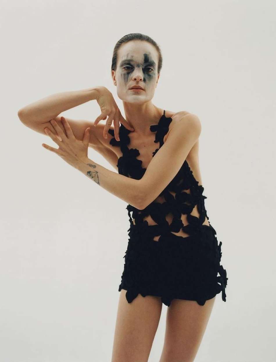 Woman with face makeup and a black outfit strikes a pose in a studio, illuminated by soft, diffused lighting against a solid white backdrop.