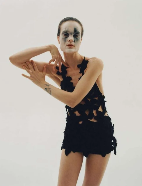 Woman with face makeup and a black outfit strikes a pose in a studio, illuminated by soft, diffused lighting against a solid white backdrop.