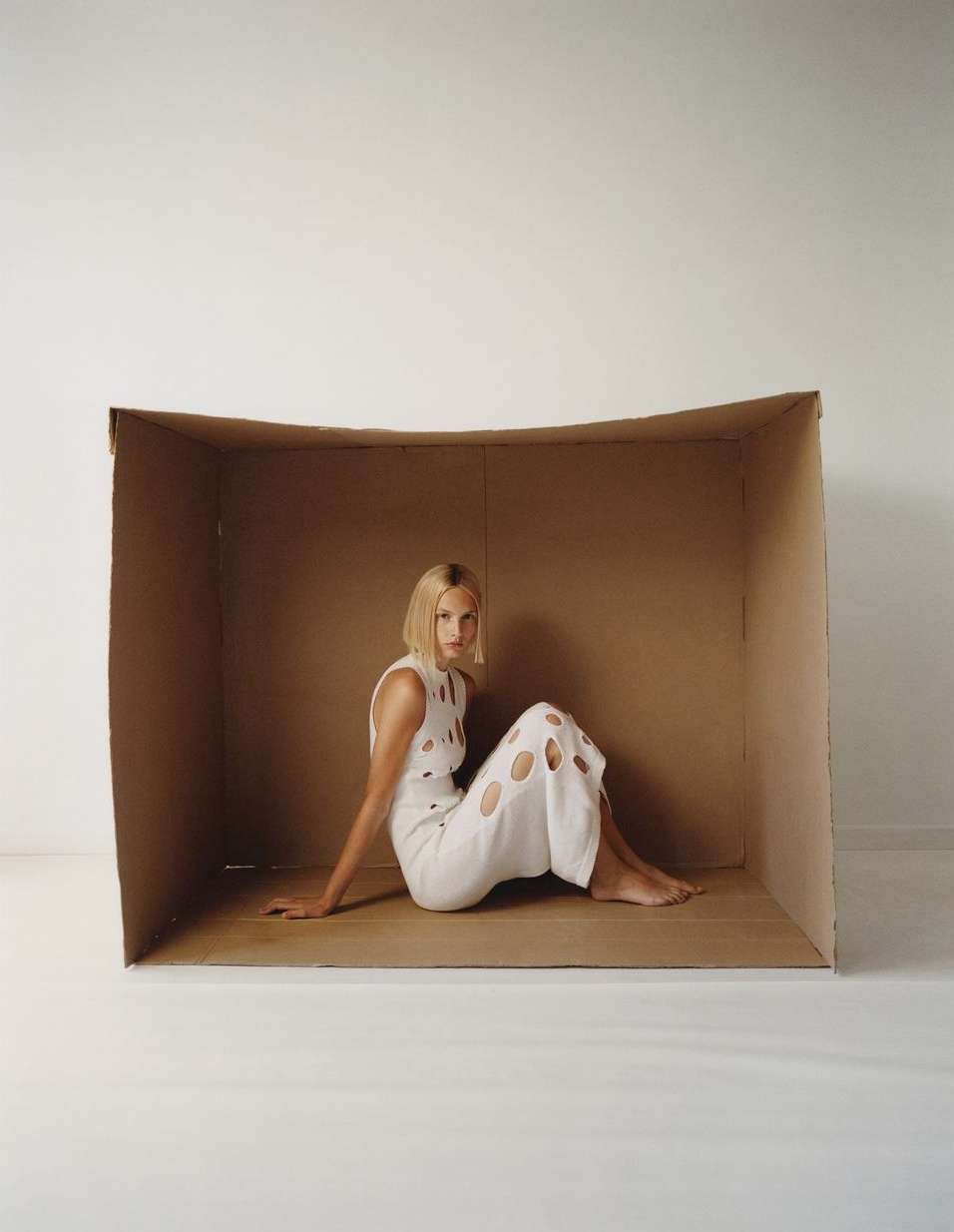Adult female model in a white cutout dress poses seated inside a cardboard box against a white backdrop in a fashion photography studio.