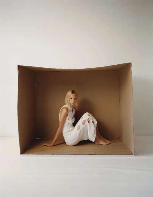 Adult female model in a white cutout dress poses seated inside a cardboard box against a white backdrop in a fashion photography studio.