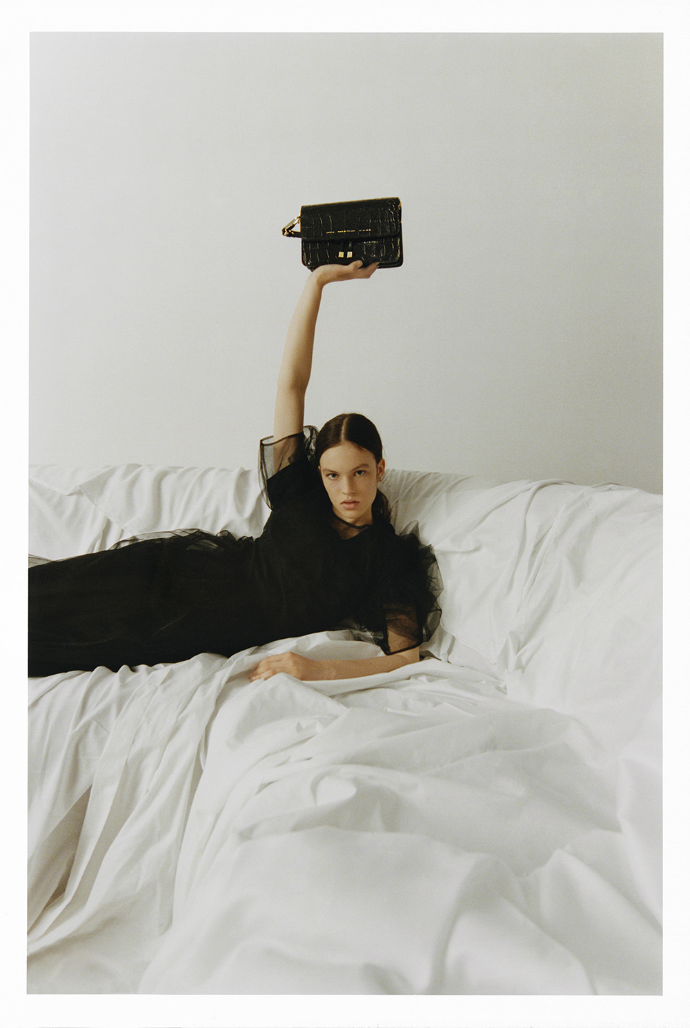 Elegant woman reclines on white sheets, holding up a black handbag, showcasing fashion and style against a minimalist backdrop.