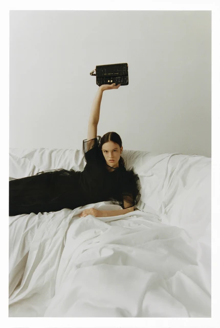 Elegant woman reclines on white sheets, holding up a black handbag, showcasing fashion and style against a minimalist backdrop.