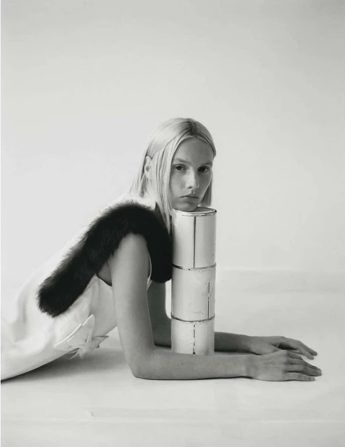 Monochrome portrait of a blonde woman in a light dress with dark fur trim, posing with a stack of metal cans in a photo studio.