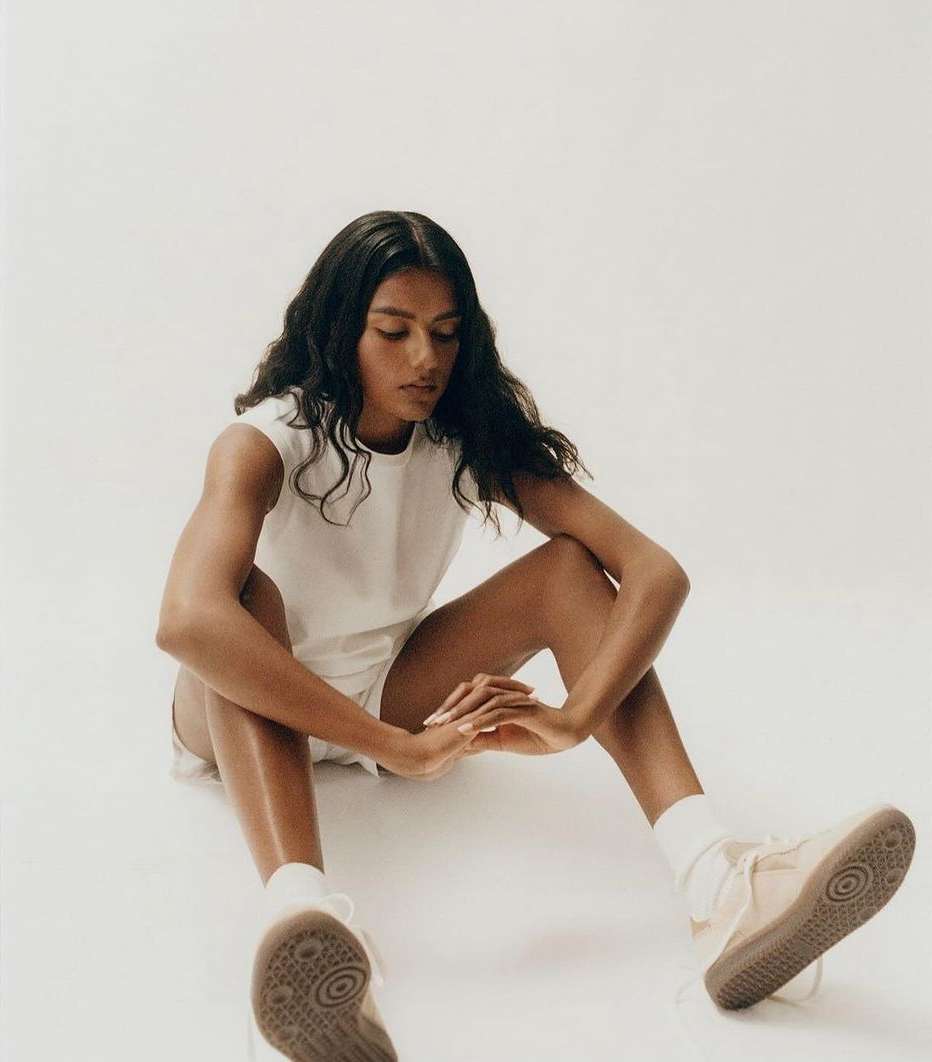 A young woman with long dark hair sits on the floor in a white shirt and shorts, wearing sneakers, in a brightly lit studio setting.